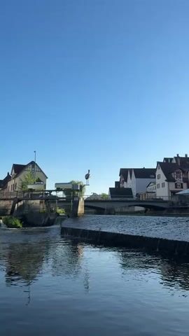 The video pans right to show a calm river in a European town under a clear blue sky.  The river's surface reflects the sky and nearby buildings.  In the distance are traditional houses with pitched roofs, some with red shutters and others with white walls.  A bridge spans the river, and a car crosses it.  A statue of a stork stands atop a concrete structure in the river.  A small waterfall is visible near the statue.  Trees and greenery dot the riverside landscape.