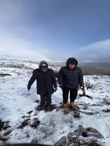 Two individuals stand in a snow-covered landscape under a partly cloudy sky with a rainbow visible in the background. Both are wearing winter clothing, including jackets and gloves, appropriate for the cold weather. The person on the left has their face partially covered, and the person on the right is wearing a hooded jacket. They raise their arms, seemingly waving or gesturing towards the camera or the surrounding scenery. The ground is covered with snow, and some sparse vegetation is visible in the snowy landscape. The setting appears to be on a high vantage point, possibly a mountain top, given the panoramic view of the surrounding terrain.