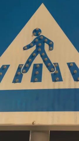 This video shows a close-up view of a pedestrian crossing sign. The sign is triangular with a blue border and a blue stripe across the bottom. Inside the triangle, a white silhouette of a person is walking across blue stripes representing a crosswalk. The silhouette is made up of small, silver studs. The studs intermittently light up with a bright blue light, creating a flashing effect. The video alternates between the sign with the studs unlit and the sign with the studs illuminated. The background is a dark blue and the sign is mounted on a reflective surface.