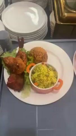 The video shows a buffet spread. There are stacks of white plates and gold-colored food warmers. Plates are arranged with a scoop of brown rice, yellow rice in small orange bowls, fried food, and lettuce. One plate includes a small white bowl of liquid and a fish. A black water bottle is also visible.