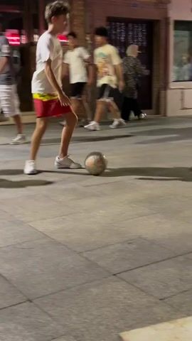 A young person in red shorts and a white t-shirt plays with a soccer ball on a paved surface at night. The person is performing tricks with the ball, kicking it with their feet. Other people are visible in the background, some walking and others standing. There is a car parked in the background and buildings on either side of the street. The lighting suggests it is nighttime or dusk. The scene appears to be a public street or walkway.