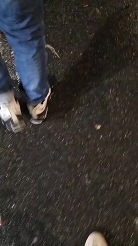 The video shows someone walking on a wet street at night. There are buildings and some sort of stalls or booths set up, possibly for an event or market. A group of men can be seen standing on the street. The environment suggests an urban area with street lighting. The scene is a bit blurry due to the camera's movement.