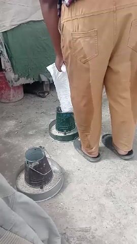A person in tan pants and black sandals is filling a green chicken feeder with food from a white bag.  Another feeder, metal and used, sits nearby. The ground is concrete. There are other items present, including a red bucket and some tarps.