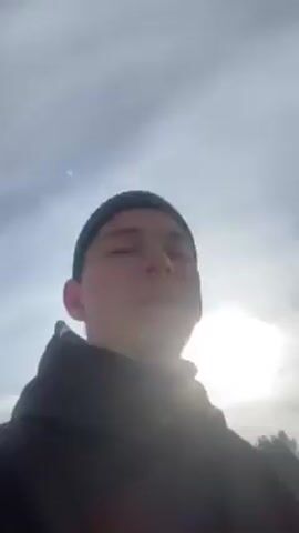 The video shows a close-up of a young man wearing a black beanie and a black jacket, standing in a snowy landscape. The sun is shining brightly behind the clouds, creating a hazy effect. The background features a gentle slope covered in snow, with a line of evergreen trees along the top of the hill. The man looks directly at the camera and appears to be speaking. The camera angle suggests the video is a selfie.