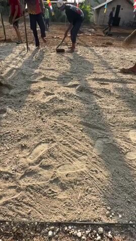 The video shows an outdoor scene with several people in what appears to be a village or rural area. The ground is covered in dirt and sand, with some footprints visible. There are several wooden poles with Indonesian flags, which are red and white, decorating the area. In the foreground, a person is holding a shovel, and another person appears to be raking or clearing the ground. Further back, more people are visible, some also holding tools like shovels. The overall atmosphere suggests some kind of community activity or preparation, possibly for an event. The sun is visible in the sky, indicating it's daytime. The background shows some simple structures, possibly houses, and trees.