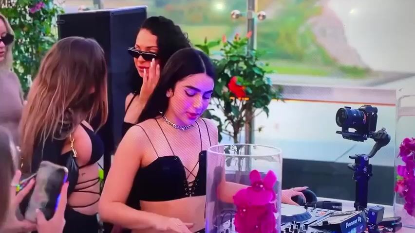 Here's a description of the video:

A woman with long dark hair, wearing a black top, is DJing at what seems to be an outdoor event. She is operating a Pioneer DJ set, which has a transparent cylinder filled with pink flowers placed on top.  A DSLR camera on a gimbal is positioned to record her as she works. 

Several other women surround the DJ booth. One is taking a picture with her smartphone. Another woman with dark hair and sunglasses stands to the side, observing the DJ.  Another woman in a tan dress and sunglasses stands in the background.

The background shows a green and white wall, suggesting an outdoor setting. Speakers are visible behind the DJ. The scene is well-lit.  The overall atmosphere suggests a stylish and social event.