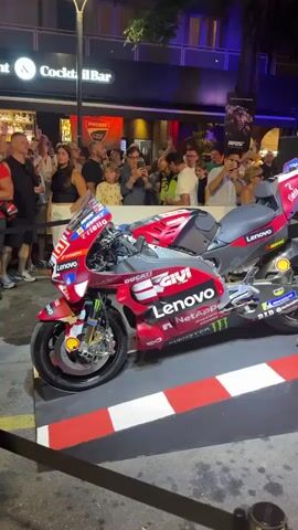The video shows a red Ducati racing motorcycle on display, with several people gathered around it. The motorcycle is positioned on a raised platform with red and white striped borders. The background features a building with a sign that reads "Cocktail Bar" and "Ducati." Many people are holding up their phones, likely to take pictures or videos of the motorcycle. The lighting suggests it is evening or night. The atmosphere appears to be a public event or exhibition.
