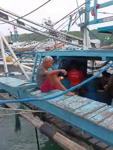 An elderly man with short, grey hair sits on a blue wooden walkway of a fishing boat, wearing a grey t-shirt and pink shorts. He is barefoot and appears to be eating something from a small white container. Behind him, inside the boat's cabin, another person in a red t-shirt is partially visible. The boat is docked with several other fishing boats in the background, suggesting a harbor or fishing village. The sky is overcast, and the water appears calm. The scene is captured from a slightly elevated angle, focusing on the man and the boat he is on.