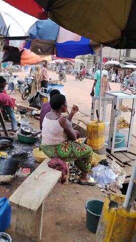 The video shows a street scene with a food vendor operating under an umbrella. A woman is sitting on a bench eating a snack. Another person is visible handing money to the food vendor. In the background, the street is bustling with people, motorcycles, and other vendors. The scene appears to be in a market or a busy area. The food vendor has a display case with food items inside. There are various containers and utensils around the vendor's stand. The ground appears dusty and cluttered.