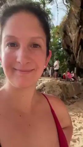 A woman in a red bathing suit stands on a sandy beach, smiling at the camera while holding it at arm's length. Behind her is a large rock formation with caves. Several people are scattered on the beach in front of the rock formation. One person is hanging from a rope inside one of the caves. Other people are sitting, standing, and milling about on the sand. There are beach towels and bags on the ground. The overall environment suggests a sunny day at a beach with rock climbing possibilities.