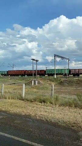 A freight train consisting of multiple boxcars and gondola cars travels along a railway track. The train is moving from left to right across the frame. The cars are a mix of red-brown and green colors. Overhead power lines and poles are visible above the train. The scene takes place outdoors on a sunny day with a blue sky and fluffy white clouds. In the foreground, there is dry grass, a dirt path, and a simple fence with white posts. Some sparse vegetation and trees are present in the midground. The overall environment appears to be rural or undeveloped.