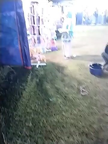 The video shows an outdoor night scene at what appears to be a carnival or fair. There's a man crouching near a bucket on the grass, while another person, wearing a light-colored outfit and a headscarf, stands nearby. There are structures and tents in the background, illuminated with lights, possibly food stalls or game booths. In the distance, there are carnival rides lit up with various colors, and people are seen walking around the fairgrounds. The ground is mostly grass. The overall atmosphere suggests a festive event taking place at night.