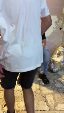 The video depicts a young man sitting on a wooden bench outside what appears to be a restaurant. He is wearing a black t-shirt with the words "JUST DO IT" printed on it and blue jeans. He is holding a cigarette in his hand. Another man, wearing a white t-shirt and black shorts, stands near him. A third man, also wearing a white t-shirt, puts on a white fedora. The restaurant's interior is visible through the glass doors, showing other patrons seated inside.