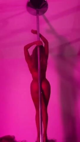 A person is performing a pole dance in a room with pink lighting. The person is wearing lingerie and high heels. They are gripping a silver pole and moving their body in a fluid motion. The background is a solid pink wall.