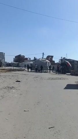 This video, likely captured in Gaza, depicts a scene in what appears to be a war-torn or severely damaged urban environment. The sky is a clear, bright blue, suggesting a sunny day. The ground is unpaved and dusty, with debris scattered around.

In the foreground, a few individuals are walking across the dusty road. In the middle ground, a group of people are gathered around a large object covered in a grey tarp. The object appears to be on wheels, possibly a cart or a makeshift vehicle. Some people are pushing or pulling this object, suggesting they are engaged in transporting it. The group consists of several men, their movements indicating a concerted effort to move the covered item.

In the background, the buildings are heavily damaged, with visible structural damage and debris. Some buildings appear to be partially destroyed or in ruins, with exposed frameworks and missing walls. The overall setting conveys a sense of destruction and displacement.

There are no clear indications of the specific time of day other than it being daylight. The weather appears to be fair and sunny. No visible text is present in the video. The activity suggests the residents are engaged in salvage, relocation, or reconstruction efforts in a challenging environment. The expressions on the faces of the people are not clearly discernible due to the video quality and distance, but their collective action implies a purpose-driven activity, likely related to survival or rebuilding amidst the devastation.