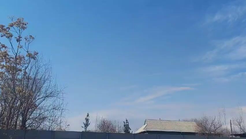 The video shows a view of the sky and rooftops on a sunny day. The sky is bright blue with thin, wispy clouds. In the foreground, there is a concrete fence.  Behind the fence are bare trees and the roofs of several buildings, including a light-colored tile roof.