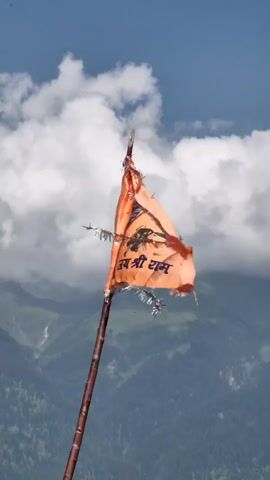 This media file features an orange prayer flag attached to a wooden pole, fluttering in the wind. The flag is tattered and appears worn, with some silver fringe remnants visible. The background depicts a majestic mountainous landscape under a partly cloudy sky, suggesting an outdoor setting. A small bird is visible in the sky. The image has a religious or spiritual feel, possibly indicating a place of worship or a sacred site. The visible text on the flag appears to be in Hindi or a similar Indian script, with the word "राम" (Ram) clearly discernible, hinting at a Hindu religious context. The lighting suggests it is daytime.
