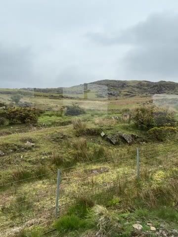 The video shows a rural landscape on a cloudy day. In the foreground, there's a wire fence and grassy terrain with some rocks. The land slopes upwards to a hill with sparse vegetation and rocky outcrops. In the distance, more hills are visible under the overcast sky. The overall impression is of a remote, unpopulated area.