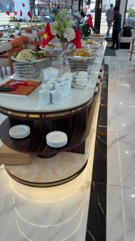 The video shows a buffet table at what appears to be a Vietnamese restaurant or event. The table is decorated with Vietnamese flags and flowers. There are various dishes on display, including salads, spring rolls, mixed vegetables, and sushi. There are also plates, bowls, and utensils available for customers to serve themselves. People can be seen in the background, some seated at tables and others near the buffet. The overall setting is bright and modern, with marble flooring.