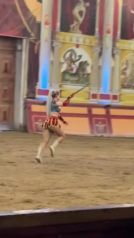 A woman with long blonde hair pulled into a ponytail, wearing a gladiator-like costume consisting of a red, blue, and gold top, a red and gold patterned skirt, and white boots, runs across a sandy arena. She is holding a sword in her right hand. In the background, a stage is decorated with Roman-themed murals and columns. Another woman, dressed similarly but with dark hair, is also visible in the background, running away from the camera. There are also some set pieces and decorations that suggest a historical or theatrical performance. The lighting is dim with spotlights illuminating parts of the stage.