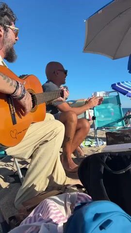 A beach scene unfolds under a clear blue sky. In the foreground, a person is partially visible, playing a guitar. To their right, a man with a shaved head sits on the sand, wearing sunglasses and clapping his hands. He has tattoos on his legs. Behind him, two women are dancing on the sand. The woman on the left wears a colorful, patterned dress, while the woman on the right wears a white bikini and a baseball cap. They are positioned in front of beach umbrellas and chairs. The sand is visible throughout the scene, with some beach bags and personal belongings scattered around. Other beachgoers and the ocean can be seen in the background.
