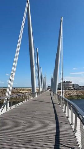 A first-person perspective shot shows a person walking across a modern pedestrian bridge on a sunny day. The bridge has a wooden walkway and sleek, white, angled support beams that rise towards a clear blue sky. The railing of the bridge is made of metal cables and posts. To the left of the bridge, there is a body of water, and beyond that, a sandy beach with some vegetation. In the distance, on the right side of the bridge, there are buildings that appear to be part of a town or city. The sun casts long shadows across the bridge, indicating it is likely morning or late afternoon. A single person is visible walking further down the bridge, away from the camera.