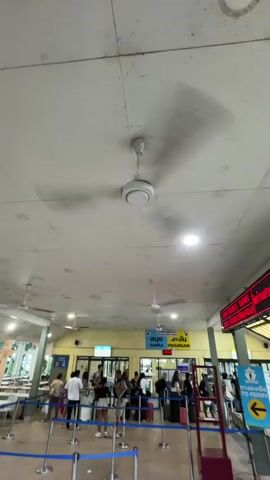 The video shows a waiting area, likely a ferry terminal, with a group of people queuing. A ceiling fan is visible at the top of the frame. The area appears to be indoors with tiled floors and a plain ceiling. People are standing in line behind blue stanchions, some with luggage. Signs are visible in the background, some with text in a foreign language and an arrow pointing to "TO FERRY". The overall atmosphere seems to be one of anticipation and movement.