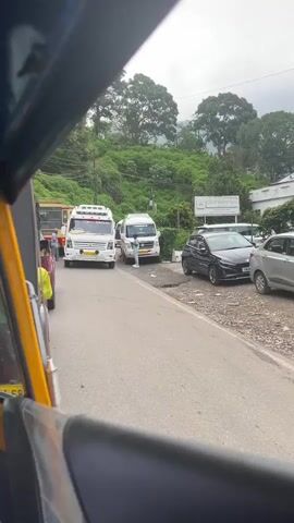 The video is filmed from the perspective of someone inside a vehicle, possibly an auto-rickshaw, given the yellow frame visible in the foreground. The vehicle is stationary on the left side of a paved road. In front of the camera, a white tempo traveler van is stopped, facing the camera. To the left of the van, a red and yellow bus is also stopped. On the right side of the road, several cars are parked, including a black hatchback and a silver car. Behind the parked cars, there's a white sign with text that is partially visible, and lush green trees and foliage cover a hillside in the background under an overcast sky. The sound of an engine is audible.