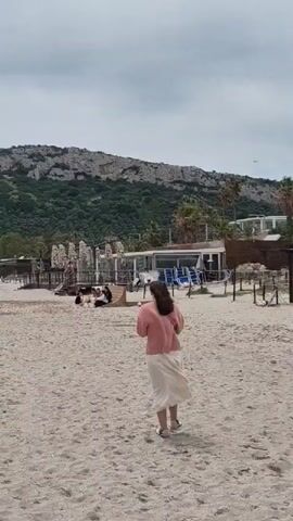 The video depicts a beach on a slightly overcast day. Initially, a woman is walking away from the camera, dressed in a pink sweater and a long white skirt. The beach is sandy with some structures and people gathered in the background. Behind the beach is a densely vegetated hill with rocky outcrops. The camera then pans to the left, showing the shoreline. The water is choppy with waves breaking on the shore, which consists of sand closer to the beach and darker, possibly pebbled or seaweed-covered areas closer to the water. In the distance, the bay is filled with sailboats and the view continues up another hilly coast.