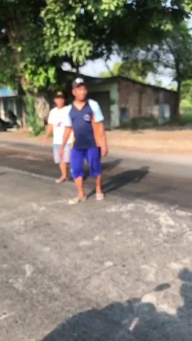 The video shows two young males standing on an asphalt road. The male in the foreground is wearing a blue and orange shirt, blue shorts, and a black cap. The male in the background is wearing a white shirt and shorts. Both appear to be looking towards the right of the frame. The environment appears to be rural or suburban, with trees and a building visible in the background. The lighting suggests it is daytime, likely late afternoon due to the long shadows. The image is somewhat blurry and the focus is on the people.