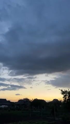 The video shows a dusky sky filled with thick, dark clouds. The sun is setting, casting a yellow-orange glow along the horizon that peeks through the lower clouds. Silhouettes of houses and trees are visible at the bottom of the frame against this backdrop. The scene is still and quiet, suggesting a peaceful, somewhat ominous evening.