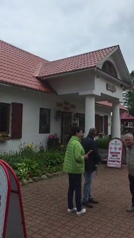 The video begins with a shot of a white building with a red tiled roof and brown shutters. There is a sign in front of the building that reads "TERRITORY OF BELARUSSIAN LANGUAGE". People are seen walking in front of the building. The video then pans to show a picturesque landscape with green fields, trees, and a body of water. Several wooden structures, possibly historical replicas or themed buildings, are visible in the distance. The camera then focuses on a path leading towards the water. Further along the path, two wooden boats resembling Viking ships are displayed. One boat is closer to the camera and is adorned with colorful shields. People are seen walking along the path towards the boats and the water. The final shot shows two individuals, a woman and a man, walking away from the camera, with trees and the water in the background. The weather appears to be overcast with some clouds and patches of blue sky.