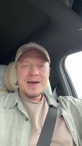 A man sits in the driver's seat of a car, wearing a tan t-shirt, a light green button-down shirt, and a tan baseball cap. He is singing and looking at the camera, moving his head slightly to the music. He wears a seatbelt. The interior of the car is visible with beige seats, a dark roof, and part of the dashboard. There are items piled up in the back seats. Outside the car, a blurry, light-colored landscape can be seen through the windshield and windows.