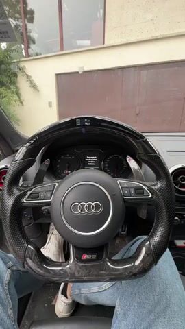 A person in a denim jacket is seated in the driver's seat of an Audi RS 3. They insert a key and turn on the car. The car dashboard lights up, and the engine starts. The steering wheel is made of carbon fiber and leather, with the Audi logo in the center and "RS 3" written at the bottom. The car is parked outdoors, in front of a house with a garage.