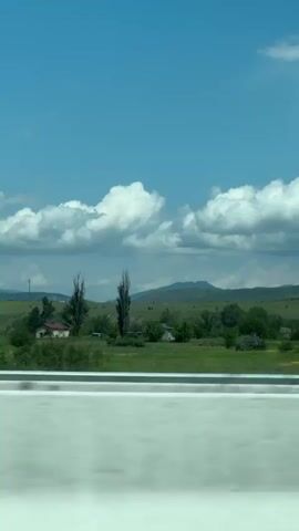 This video shows a landscape from a moving vehicle. The scene is dominated by a bright blue sky with scattered white clouds. Below the sky is a green landscape with rolling hills, trees, and some vegetation. In the foreground, there is a concrete barrier that appears to be part of a highway or road. The perspective suggests the video is taken from a passenger seat or the driver's side of a car. The movement of the vehicle causes a slight blur in the foreground, especially on the concrete barrier, indicating motion. The overall impression is of a journey through a rural or semi-rural area on a clear, possibly sunny day. There are no people or distinct human activities visible in the frame.