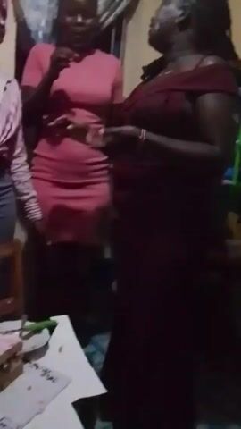 In a dimly lit room, several people are gathered around what appears to be a birthday celebration. Two women stand out in the initial frame, one in a pink dress and the other in a maroon off-shoulder dress. They seem to be talking and gesturing. The camera then pans down to reveal a table with a partially eaten cake decorated with pink frosting. A wooden chair sits nearby. Other individuals are also present, with one wearing a red and white checked shirt, and another in ripped jeans and a white shirt with a rainbow heart. The setting seems informal, suggesting a private gathering.