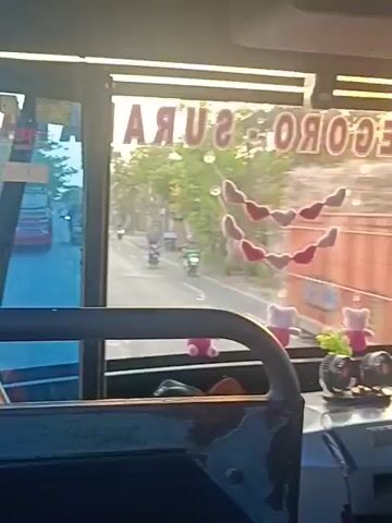 The video is filmed from inside a vehicle, likely a bus, looking out through the front windshield and side window. Through the windshield, a road is visible with traffic, including cars and motorcycles. Trees and buildings line the road. Sunlight streams in, suggesting it is daytime. A sign with Indonesian text, possibly indicating a destination or route, is visible at the top of the windshield. The side window shows more of the road and passing scenery. Inside the vehicle, parts of the dashboard and a passenger's head are visible. There are decorative items inside the vehicle, including heart-shaped ornaments and stuffed toys. The video appears to be shaky, suggesting movement of the vehicle.