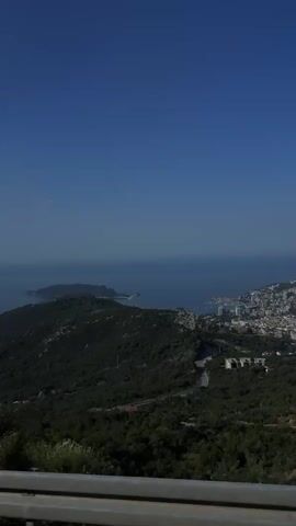This media file is a landscape video, likely shot from a high vantage point overlooking a coastal town and the sea. The dominant subjects are the natural landscape and a man-made element.

In the foreground, a circular speed limit sign with the number "60" in black on a white background, within a red border, is prominently displayed. This suggests the scene is at a road, likely on a mountain pass given the elevated perspective. To the right of the sign, there's a metal guardrail.

Beyond the immediate foreground, the scene opens up to a vast expanse of green, rolling hills densely covered with trees and vegetation. The hills descend towards the coast. In the distance, a town is visible nestled along the coastline, with buildings clustered together and stretching out towards the sea. The deep blue of the ocean stretches out to the horizon, meeting a clear, bright blue sky. The sunlight suggests it's daytime, likely late morning or early afternoon, and the weather appears to be clear and sunny.

The overall mood is serene and expansive, capturing the beauty of a coastal mountainous region. The presence of the speed limit sign and the road imply a journey or a viewpoint along a route. The town in the distance and the sea hint at a Mediterranean or Adriatic location.

Specific location cues confirm this as Lapčići, Montenegro, a known viewpoint offering panoramic vistas of the coastline and the Adriatic Sea.