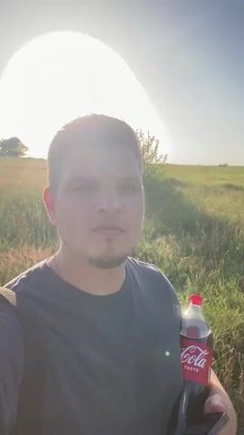 A man is holding a bottle of Coca-Cola and a phone in front of a grassy field during sunset. The man is speaking and smiling at the camera, and the sun is shining brightly in the background.