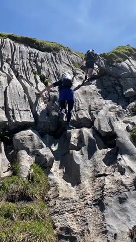 The video shows two individuals hiking up a steep, rocky mountainside. The person in the foreground, wearing a backpack and blue shorts, is actively climbing a section of the rock face, using their hands and feet to ascend. They are positioned on a ledge with a red and white marker visible on the rock to their left. Behind them, another person, also with a backpack, is further up the mountain, also climbing. The environment is sunny with a clear blue sky and patches of green grass growing on the slopes. The rock formations are prominent, with visible textures and crevices.