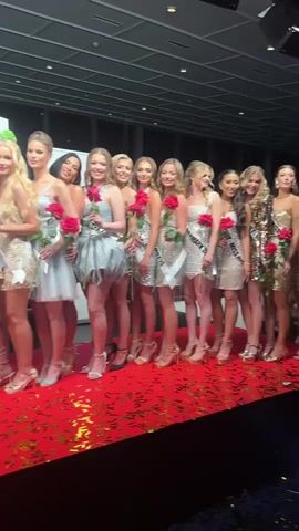 A beauty pageant appears to be taking place. A group of roughly 15 young women are posing together on a red carpet. Many are wearing short, sparkly dresses in golds and silvers, with sashes bearing words like "Miss 2023" and "Miss Rauma." Each woman holds a single red rose. Confetti litters the carpet. Later in the video, two women dressed in formal ballgowns – one red, one silver – join the group, with one woman holding a large bouquet of white and red roses. In the background is a black ceiling with recessed lighting, and a floral arrangement on a table.