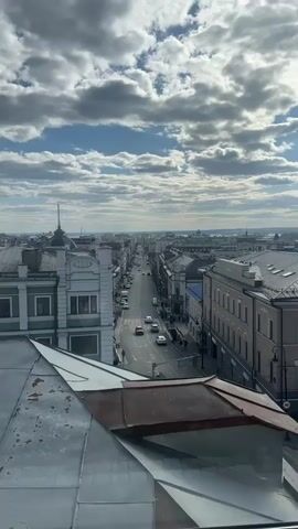 From a high vantage point, likely a rooftop, the video presents a view of a city street stretching into the distance. The street is lined with buildings of varied heights and architectural styles, mostly in light colors, and appears relatively straight, fading slightly into the horizon. Cars can be seen moving along the street, and pedestrians walk on the sidewalks. The foreground includes the edge of the metal rooftop, possibly a barrier, from where the video is shot. The sky is partly cloudy with patches of blue visible, the sunlight illuminating the scene and casting shadows from the clouds.