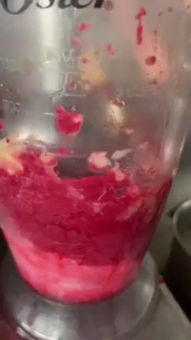 The camera captures a close-up view of an Oster blender, filled with a pink and red mixture, in operation. The mixture is being blended, creating visible splashes and movement within the container. Markings for measurement, including "3 Cups", are visible on the blender's clear plastic pitcher. The background includes glimpses of what appears to be a stainless steel kitchen counter and a pot. The focus is on the process of blending the colorful mixture, with no faces or identifiable individuals visible.