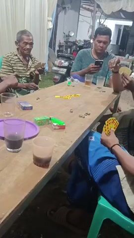 Four men are seated around a light brown wooden table outdoors. The man on the far left wears a striped shirt and holds playing cards. The man next to him, wearing a green t-shirt, is looking at his phone. In the foreground, a man in a blue shirt holds playing cards. Another man is partially visible on the right, also holding cards. The table has several glasses, some with beverages, and small boxes of cigarettes. In the background, there is a white tent structure, a motorcycle, and various household items.
