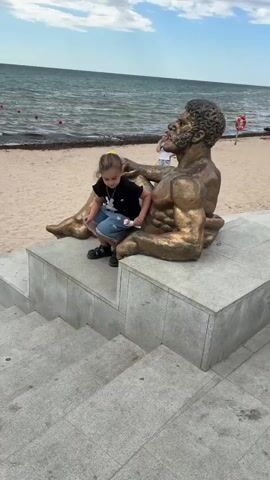 A young girl with blonde hair in pigtails sits on a bronze sculpture of a man on a concrete platform. She is wearing a black t-shirt and blue shorts. She is holding a sheet of stickers and an ice cream cone. The sculpture is seated and facing to the right. In the background, there is a sandy beach and the ocean. The sky is overcast. Another young person with blonde hair is walking in the background on the beach.
