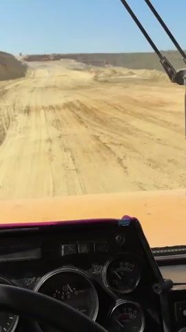 This is a first-person view from the inside of a vehicle, likely a truck, looking out through the windshield. The vehicle is on a dirt road in what appears to be a construction or mining site. Tire tracks are visible on the sandy, uneven ground. In the distance, there is a hill or embankment with more sandy terrain and some white material, possibly cement or building materials. The sky is clear and blue. The dashboard of the vehicle is partially visible at the bottom of the frame, showing a steering wheel and various gauges. A windshield wiper is also prominent in the upper right portion of the windshield. The overall impression is of a vehicle moving through an industrial or developmental landscape.