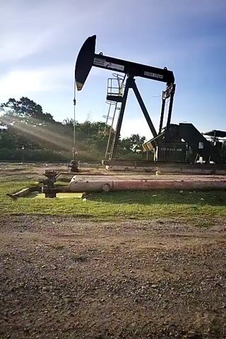 A black oil pump jack is operating in a dry, dusty field under a bright sky with some trees in the background. Sunbeams are visible in the air. The pump jack is moving up and down. There are pipes and some equipment on the ground near the base of the pump jack. There is a person lying down near the pipes.