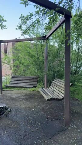 The video displays an outdoor scene featuring a weathered wooden swing set with two swings. One swing is in the foreground, angled towards the right, and appears to be slightly tilted. The second swing is further back and to the left, and is mostly stationary. The swings are suspended from a metal frame, which is partially obscured by green leaves and branches from an overhead tree. The ground beneath the swings is dark and appears to be a mixture of asphalt or packed dirt, with some wet patches and debris. In the background, there is lush green foliage and what appears to be a building structure, possibly made of brick. The overall environment suggests a park or a backyard setting during daylight hours.