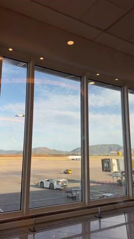 The video shows an airplane taking off from an airport. The camera is positioned inside the airport terminal, looking out through a large window. Outside, on the tarmac, there are airport vehicles and another airplane in the distance. The plane taking off is white with blue and green markings. The sky is blue with white clouds. The video ends as the plane continues to climb into the sky.