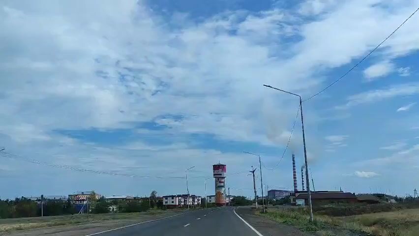 The video shows a view from a car driving on a road towards a town. On the left side of the road, there are some buildings and a water tower with colorful murals. On the right side of the road, there are some industrial chimneys emitting smoke. The sky is blue with white clouds. The road is asphalt and has white lines marking the lanes. There are streetlights and power lines along the road. The overall scene is of a rural or industrial town on a cloudy day.
